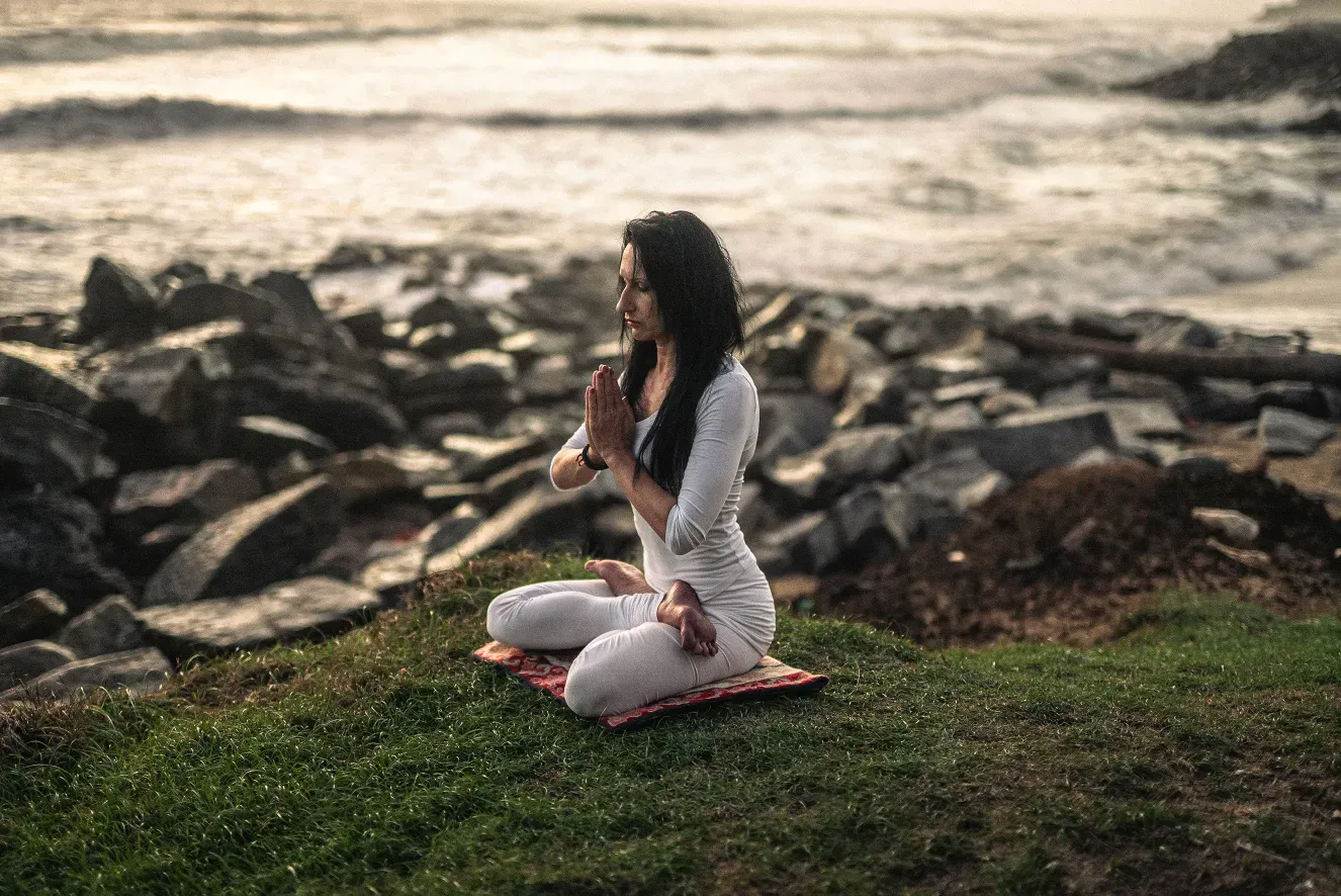 Žena v bielom sedí so zopnutými rukami v meditačnej polohe na tráve pri skalnatom morskom pobreží