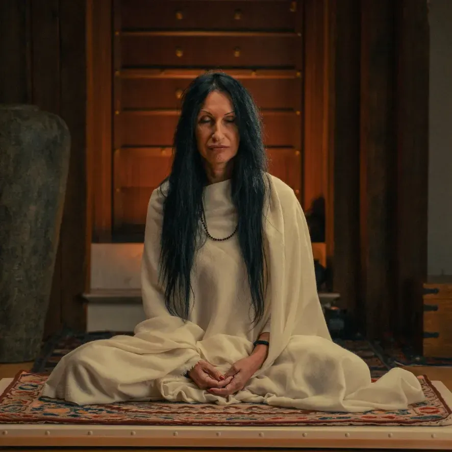 Žena v bielom odeve sedí so zavretými očami v meditácii na koberci v ášrame