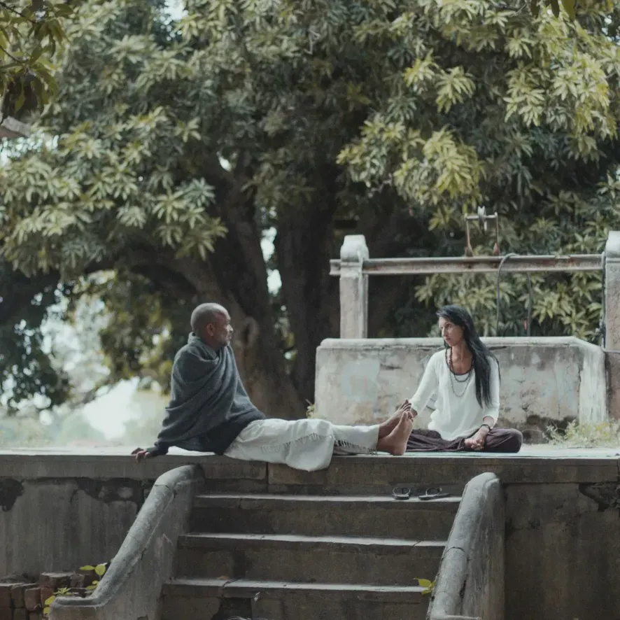 Two people sitting on a stone terrace beneath a large tree, in conversation